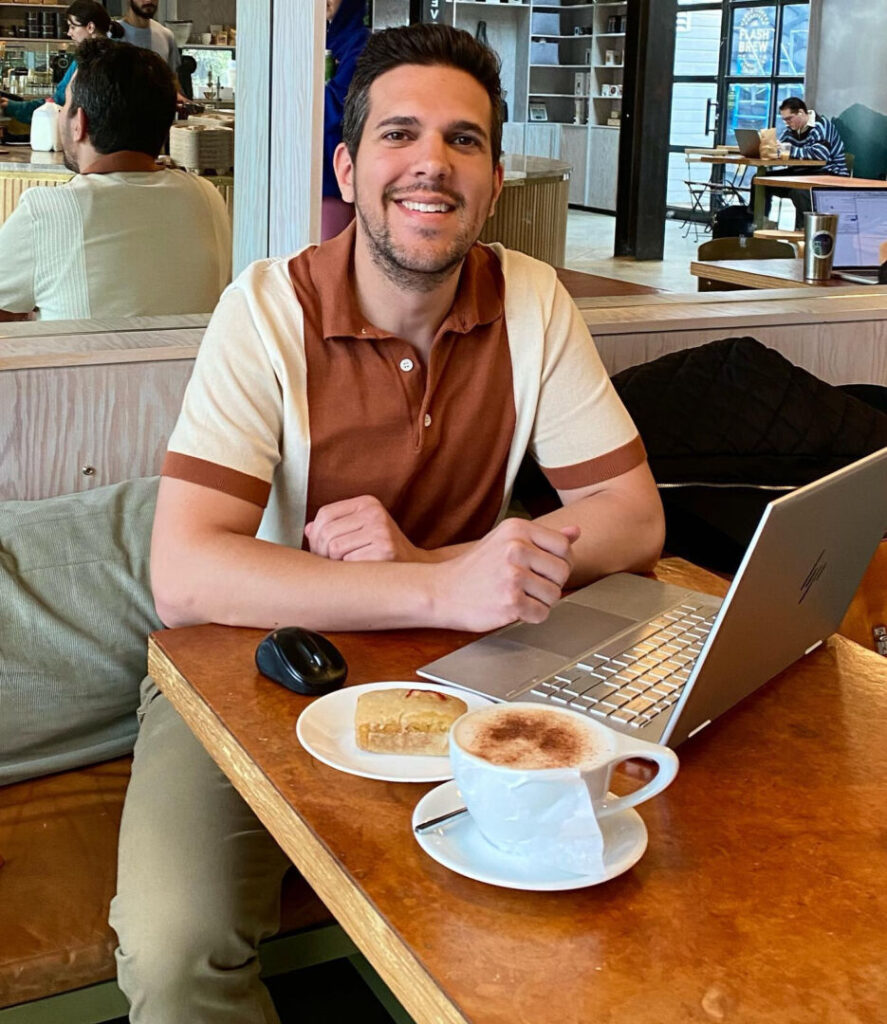Max DiNatale at a cafe and working on his laptop, a coffee cup and dessert on the table